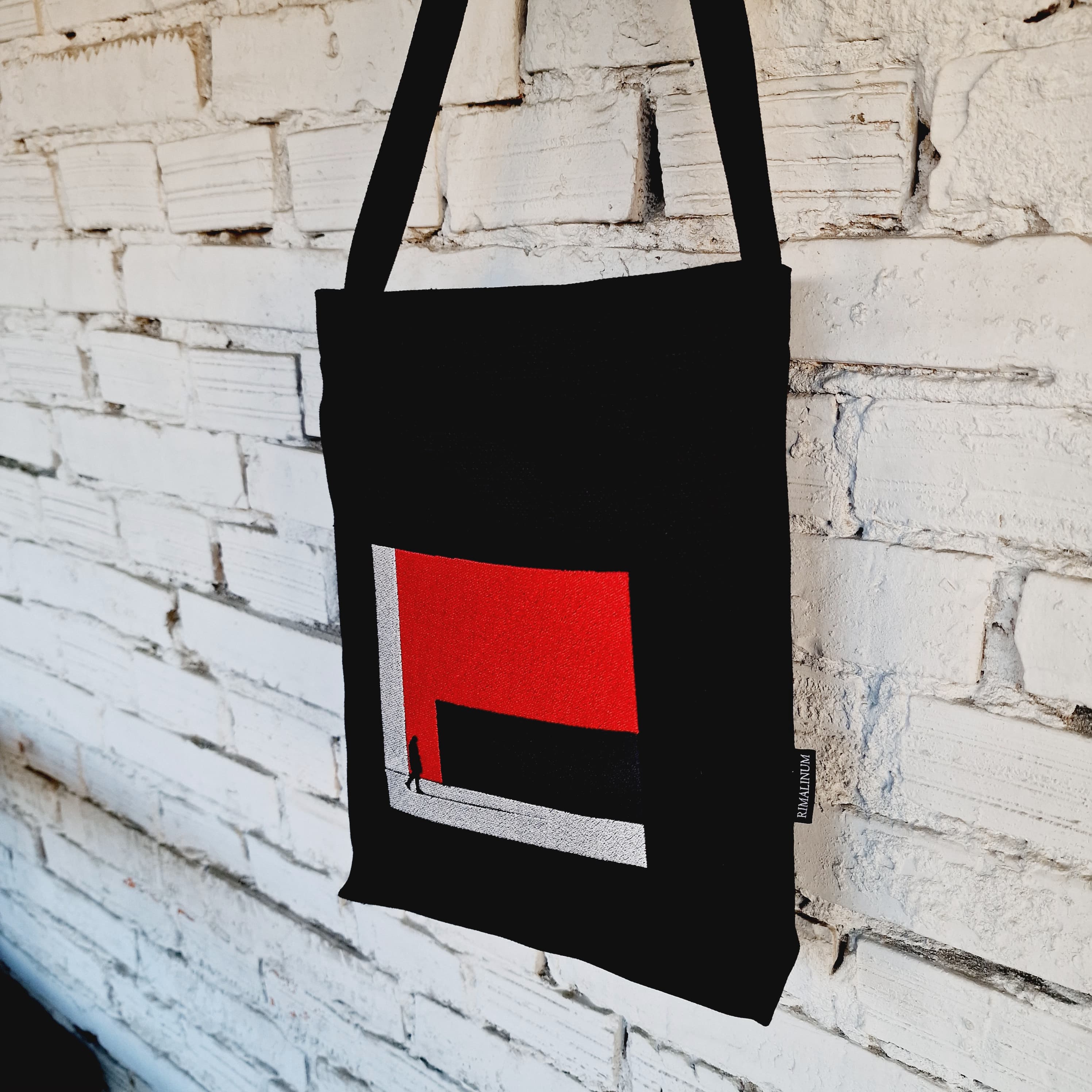 Black crossbody bag, featuring a bold red and black embroidered design, hanging on a white brick wall