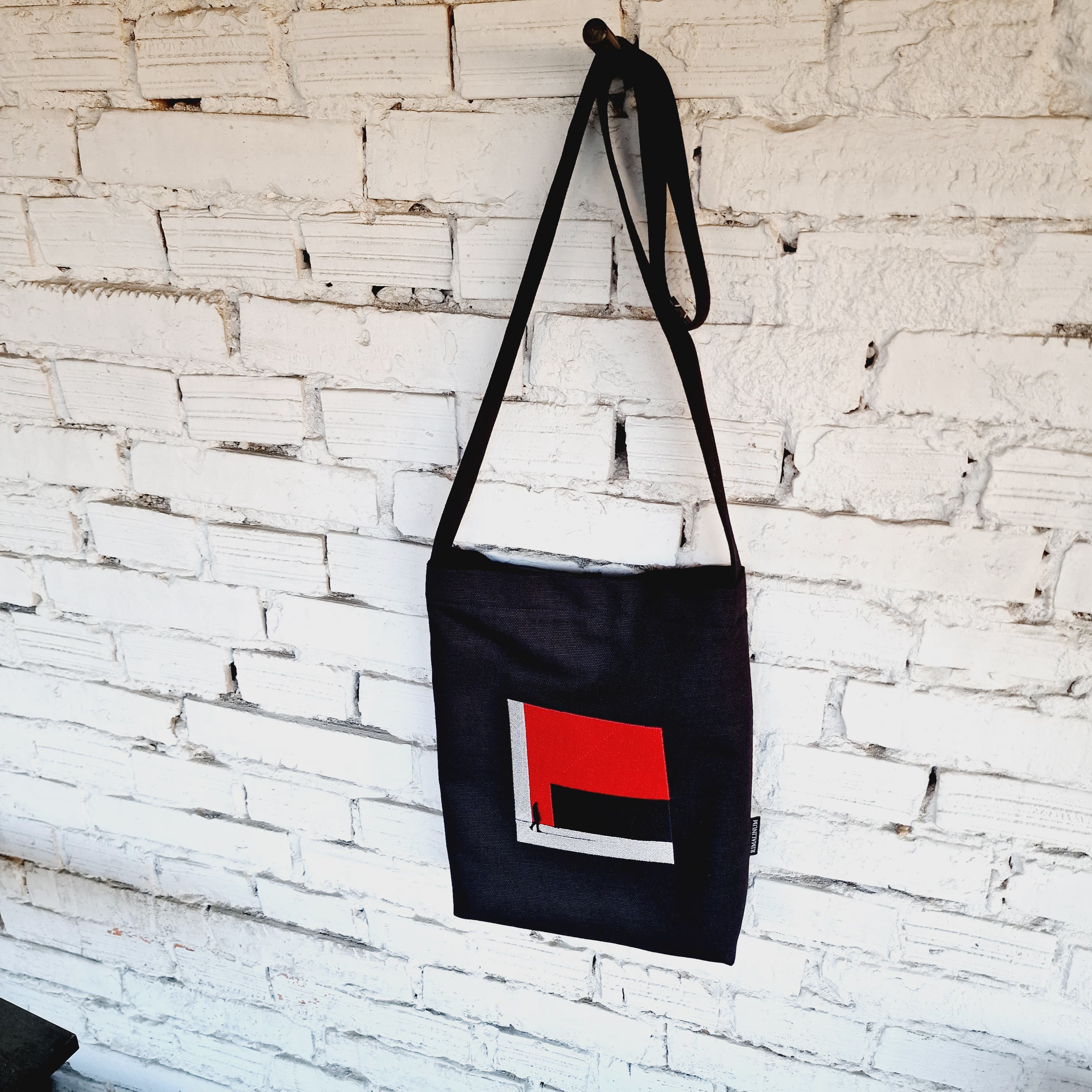 Stylish black crossbody bag with long strap and red embroidered design against a white brick wall