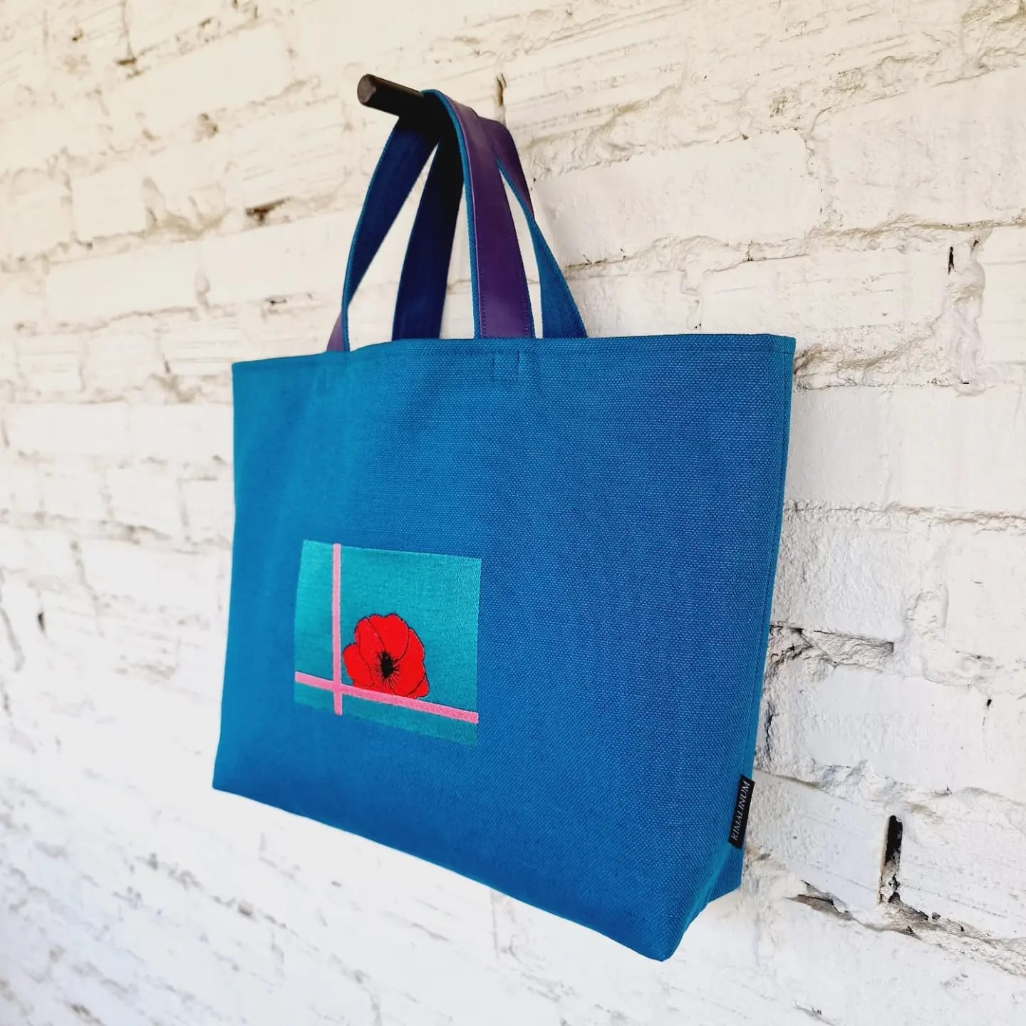 Side view of blue handbag with purple handles and artistic red poppy embroidery, hanging against a white brick wall